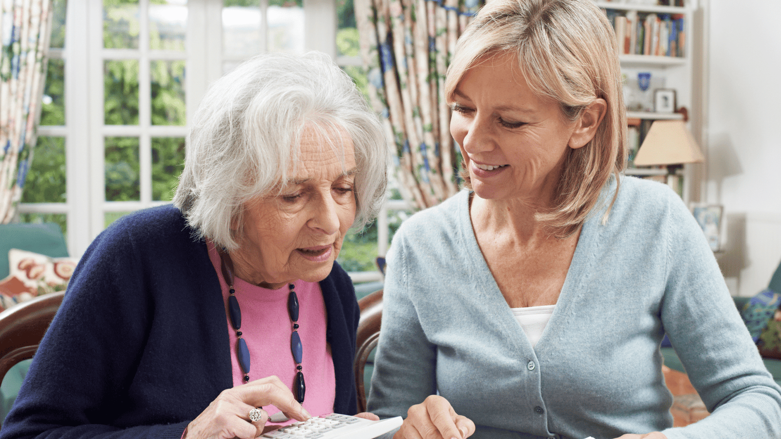 Palliative Care - An elderly woman looking focused while using a calculator and being assisted by a younger adult woman.