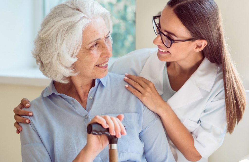 An elderly woman holding a cane, smiling at a staff member, who is smiling and has her arms on her shoulders.